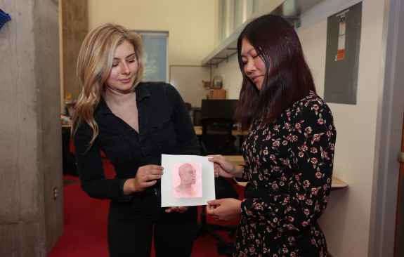 MIT graduate student researchers Paris Myers (left) and Ticha Sethapakdi watch as a drawing of a human face turns its head to the right. Thermochromorph combines CMYK imaging, laser cutting, manual printmaking, and thermochromic inks to transform images. Credits: Image: Design by Alex Shipps, photograph by Mike Grimmett/MIT CSAIL