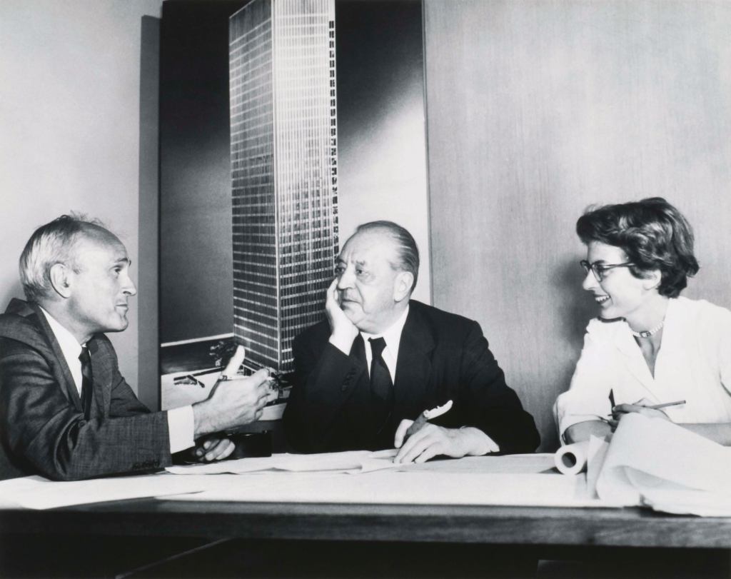 Philip Johnson, Ludwig Mies van Der Rohe, and Phyllis Lambert discuss the Seagram in front of an image of the building model soon after she helped the architects win the commission in 1955.